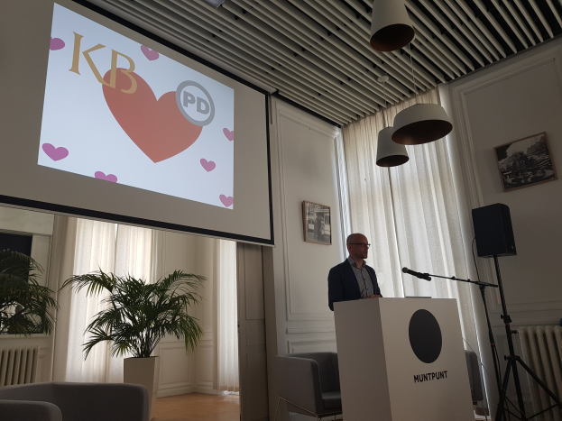 Ein Mann steht an einem Podium mit einem Mikrofon und hält eine Präsentation zum Thema Geschlechtergerechtigkeit, mit einem Projektionsschirm, einem Lautsprecher, Stühlen, einer Topfpflanze, gerahmten Fotos an der Wand, Vorhängen und Deckenleuchten im Raum.