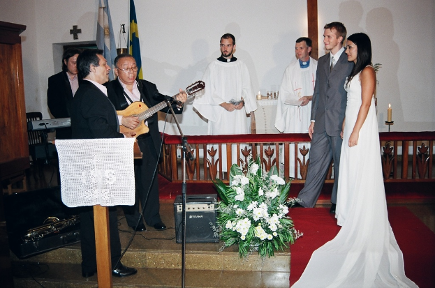 Eine Hochzeitszene in einem Raum mit zwei Pastoren, einer Braut, einem Bräutigam und drei Musikern; ein Podium mit Stoff vor den Musikern; ein Tisch mit Kerzen hinter den Pastoren; ein Kerzenhalter hinter dem Paar; ein Bukett, Lautsprecher, Taschen und roter Teppich auf dem Boden; sowie Flaggen, eine Wand mit einem Kreuz, einen Schrank und einen Stuhl im Hintergrund.