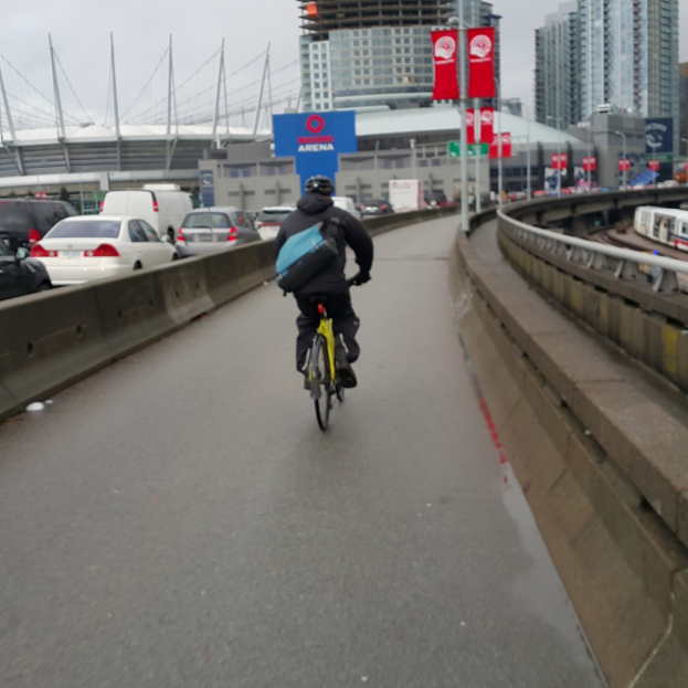 Eine Person mit Helm und Rucksack sitzt auf einem Fahrrad auf einer Straße mit parkenden Autos und Gebäuden im Hintergrund, mit Bannern an Pfählen in der Nähe.