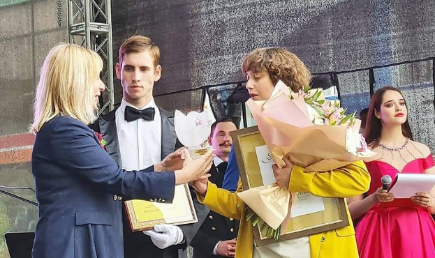 Eine Gruppe von Menschen auf einer Bühne, wobei einer in einem gelben Kleid Blumen hält und ein anderer Papiere und ein Mikrofon, während sie einen Preis erhalten.