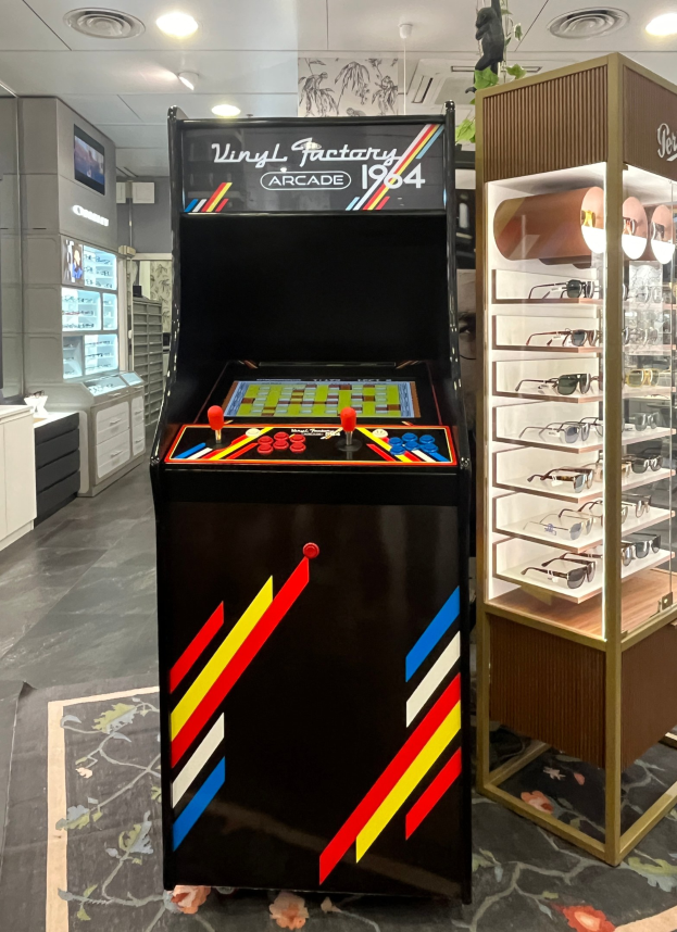 Ein Vintage-Arcade-Automat in der Mitte eines retro-stylischen Ladens mit Gläsern auf einem Gestell rechts, Schränken, einem Fernseher und Deckenlampen im Hintergrund.