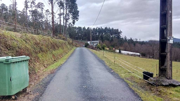 Grüner Müllcontainer am Straßenrand mit Gras, Pflanzen, Bäumen, Pfählen, Drähten und einem Zaun, unter einem bewölkten Himmel, mit einer Pfostenanzeige auf der rechten Seite und verstreuten Gegenständen auf der linken Seite.
