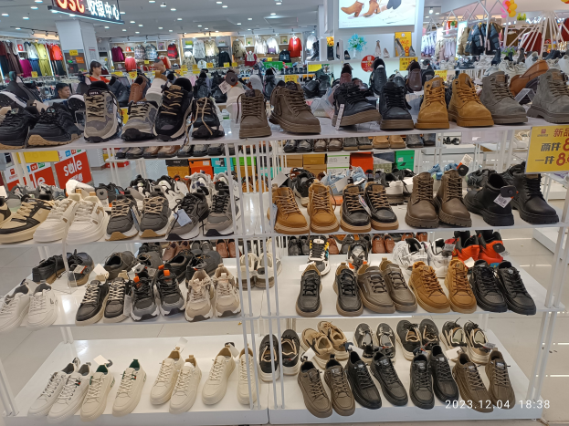 Ein Schuhgeschäfts-Innenraum mit verschiedenen Schuhen auf Gestellen, Preisschildern, Menschen im Hintergrund, hängenden Kleidungsstücken, Deckenlampen, einem Bildschirm und Texttafeln, mit einem Wasserzeichen, das einen Laden in China anzeigt.