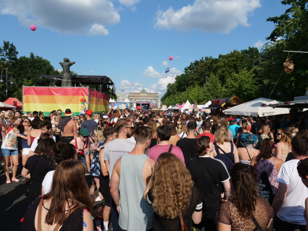 Eine große Menschenmenge, die eine Straße mit Zelten, Bäumen, Pfählen, Laternen und einer Statue gesäumt von Gebäuden im Hintergrund unter einem bewölkten Himmel mit Ballons während des Christopher Street Day in Berlin geht.