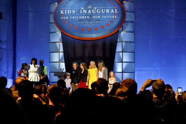 Eine Gruppe von Menschen auf einer Bühne, einige halten Mobiltelefone, mit einem Vorhang und einer Tafel mit Text im Hintergrund, was auf ein Kinderweihefest hinweist.