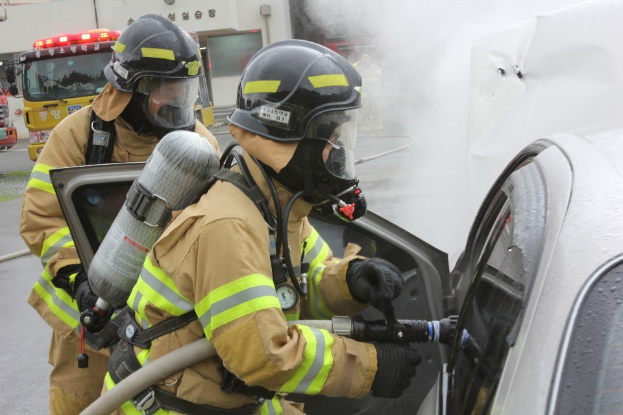 Zwei Feuerwehrleute in Schutzausrüstung verwenden einen Schlauch, um ein brennendes Auto zu löschen, mit Rauch und anderen Fahrzeugen und einem Gebäude im Hintergrund.