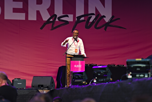 Mann in weißem Hemd und braunen Hosen, der in ein Mikrofon auf einer Bühne spricht, mit einem pinken "Berlin as Fuck"-Plakat dahinter.