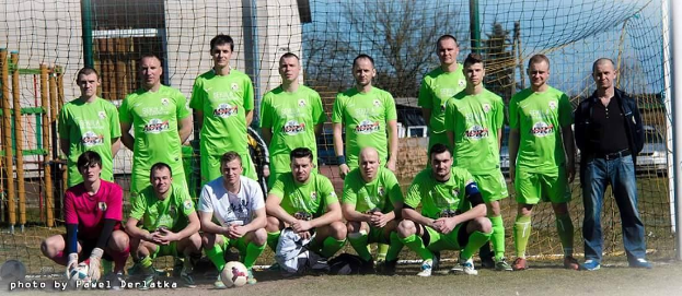 Gruppe von Männern in grünen T-Shirts auf einem Fußballfeld mit einem Ball davor, einige in Hockstellung, neben einem Torpfosten mit Bäumen, Gebäuden und Fahrzeugen im Hintergrund.