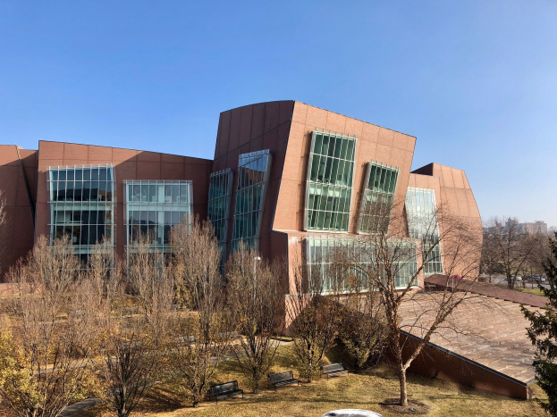 Großes modernes Gebäude mit zahlreichen Fenstern, umgeben von Bäumen, Bänken, Gras und einem Lichtmast, mit anderen Gebäuden und einem klaren blauen Himmel im Hintergrund, identifiziert als der neue Campus der University of Illinois at Chicago.
