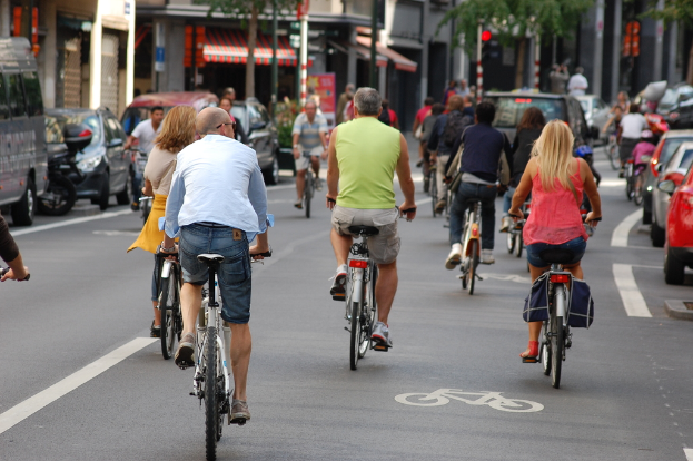 Menschen, die auf Straßen mit Autos auf der linken und rechten Seite Fahrrad fahren, und Geschäfte, ein Zelt, Bäume und Gebäude im Hintergrund.