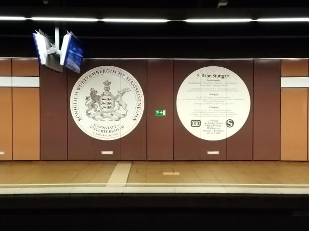 Ein Bahnsteig mit einem Schild, das 'S-Bahn Stuttgart' und ein Logo an der Wand zeigt, sowie einige Lampen an der Decke und Gegenstände auf der linken Seite.