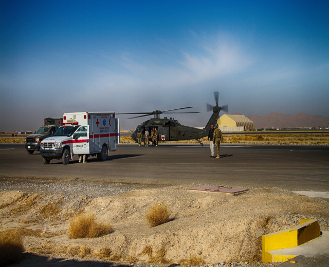 Hubschrauber auf Gras neben einem Krankenwagen mit Menschen in der Nähe, Hügel und klarer blauer Himmel im Hintergrund.