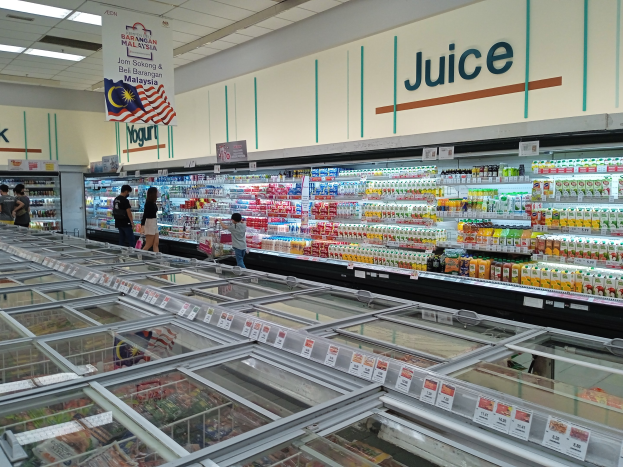 Ein gut sortierter Lebensmittelgang mit Regalen voller Getränke, frischem Gemüse und anderen Lebensmitteln, einige Kunden stöbern und eine Deckenleuchte, die den Raum erhellt.