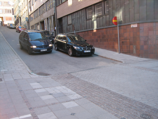 Mehrere Autos parken auf der rechten Seite einer Straße, mit Hochhausgebäuden mit Fenstern im Hintergrund.