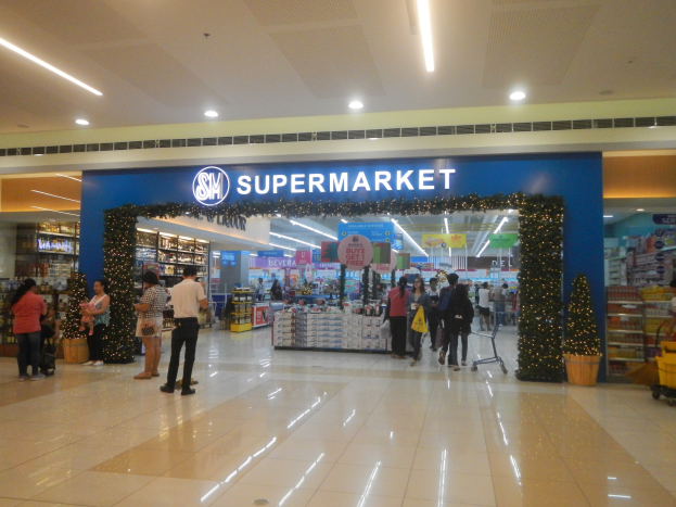 Eine Gruppe von Menschen vor einem Supermarkt, einige halten Einkaufst├Ąsche, mit einem Einkaufswagen, Topfpflanze, Regalen, Schildern und Deckenbeleuchtung.