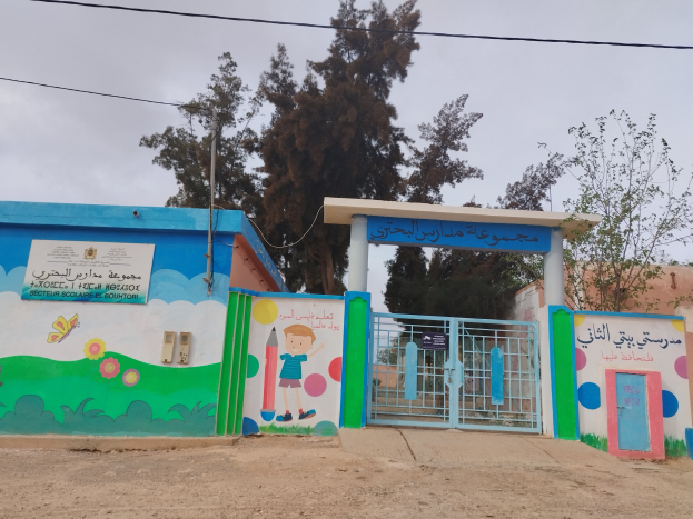 Blau-weißes Gebäude mit einer Wandmalerei, einem Tor, einer Namensschilder, Häusern, Bäumen, einem Strommast mit Drähten und einem bewölkten Himmel, das den Eingang zu einer Kinder-Schule markiert.