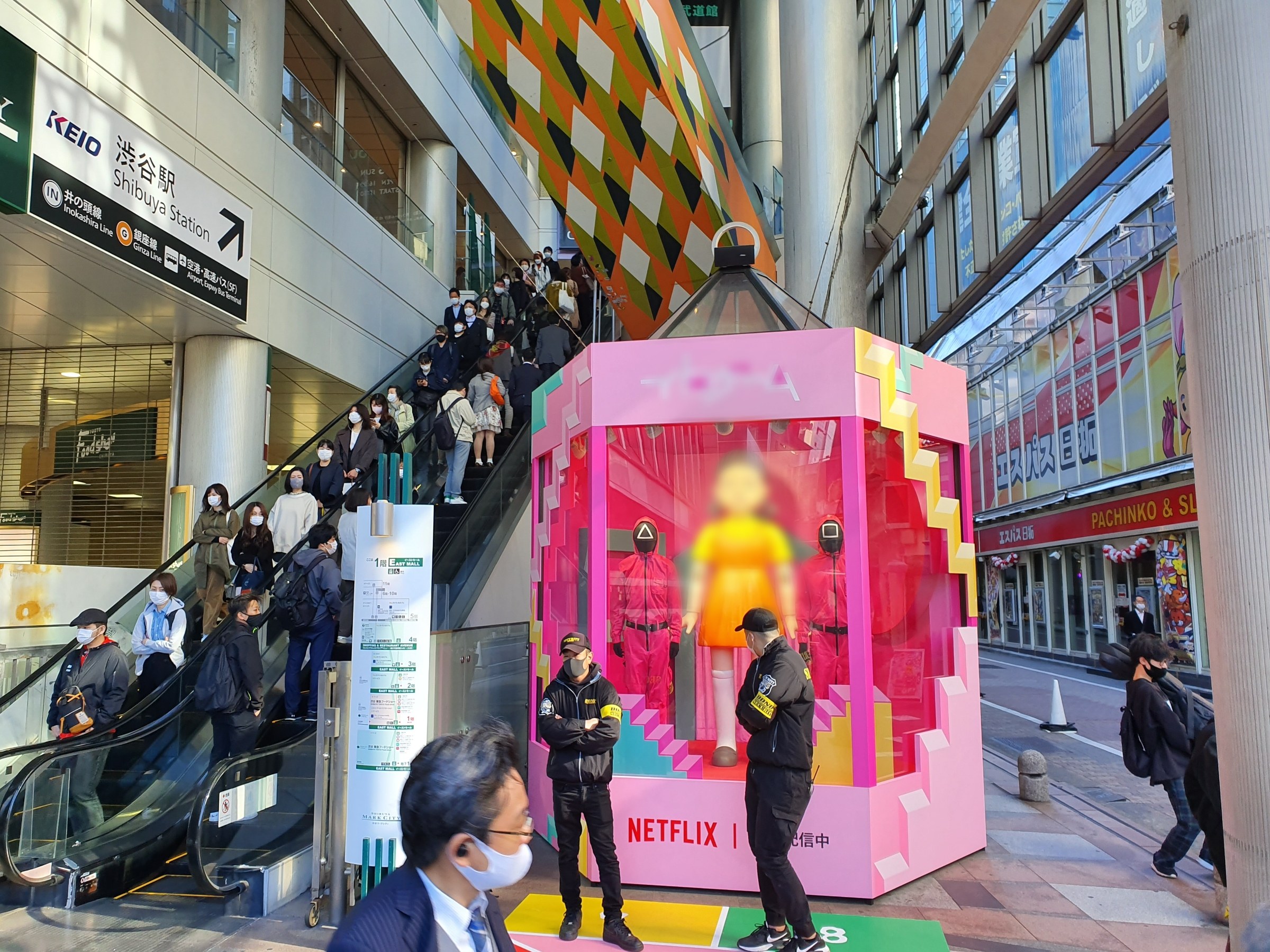 Eine Gruppe von Menschen steht vor einem Gebäude mit Glaswänden und Texttafeln, einige tragen Masken, in der Nähe einer Rolltreppe; ein pinker Kasten in der Mitte zeigt Schaufensterpuppen und trägt die Aufschrift "Netflix's new Netflix Booth in Tokyo."