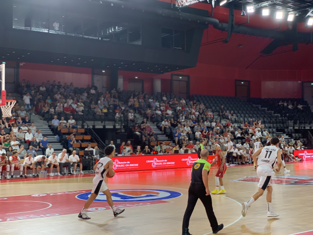 Eine Gruppe von Menschen, die Basketball in einer Turnhalle mit einem Netz auf der linken Seite spielen, Zuschauern auf Stühlen und Deckenbeleuchtung, die eine Stadionatmosphäre suggeriert.