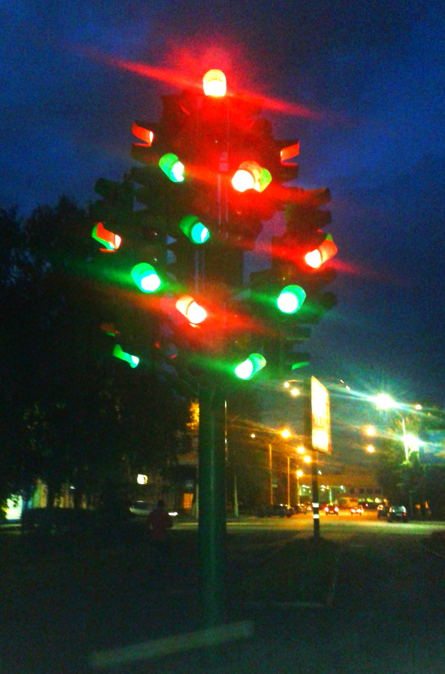 Eine nächtliche Ampel mit grünen und roten Lichtern, umgeben von Fahrzeugen, Bäumen, Gebäuden und Lichtern im Hintergrund, mit dem Himmel darüber.