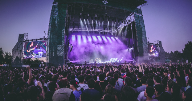 Eine große Menschenmenge schaut bei einem Musikfestival auf einer hell erleuchteten Bühne mit Instrumenten und Bildschirmen, die vor einem Gebäude, Bäumen und einem klaren blauen Himmel aufgebaut sind.