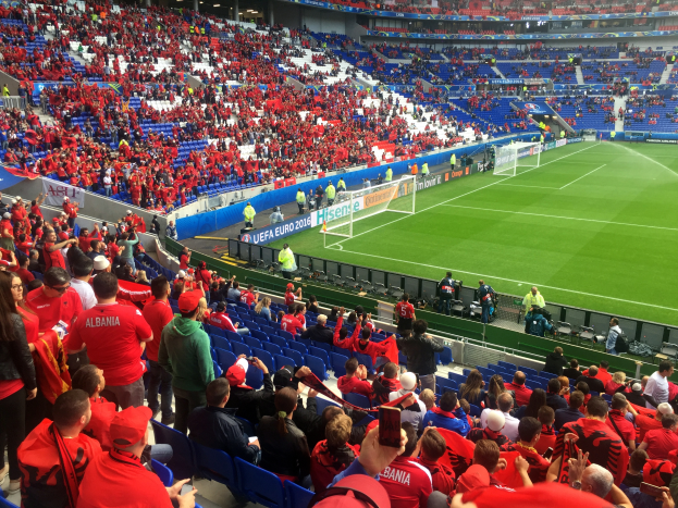 Ein volles Stadion mit Zuschauern, einige sitzen und einige stehen, mit Bannern, einem Torpfosten und Deckenleuchten, die ungeduldig auf den Beginn eines Fußballspiels warten.