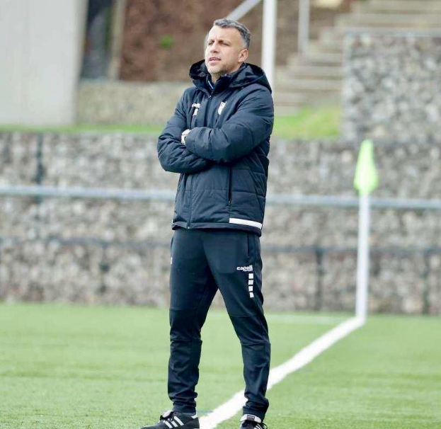 Ein Mann steht auf einem Fußballfeld mit verschränkten Armen und trägt eine schwarze Jacke und Hose, mit einem Zaun, einer Treppe und einer Wand im Hintergrund.