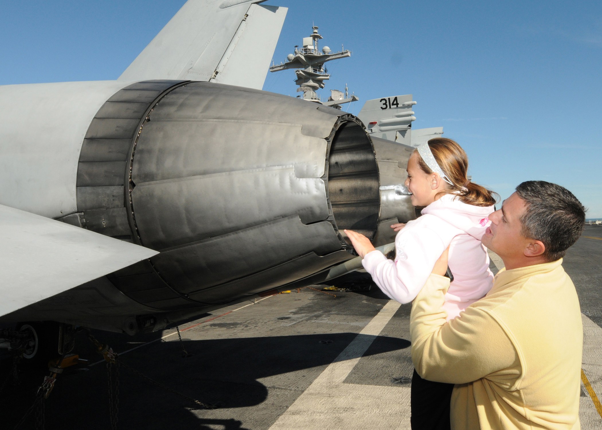Ein Mann in einem gelben Hemd und schwarzen Hosen steht neben einer Frau, die ein Baby hält, auf dem Flugdeck eines Flugzeugträgers, mit anderen Jets und einem klaren blauen Himmel im Hintergrund.