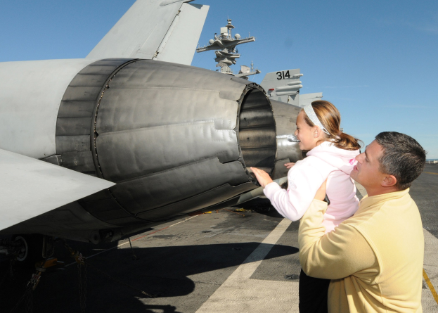 Ein Mann in einem gelben Hemd und schwarzen Hosen steht neben einer Frau, die ein Baby hält, auf dem Flugdeck eines Flugzeugträgers, mit anderen Jets und einem klaren blauen Himmel im Hintergrund.