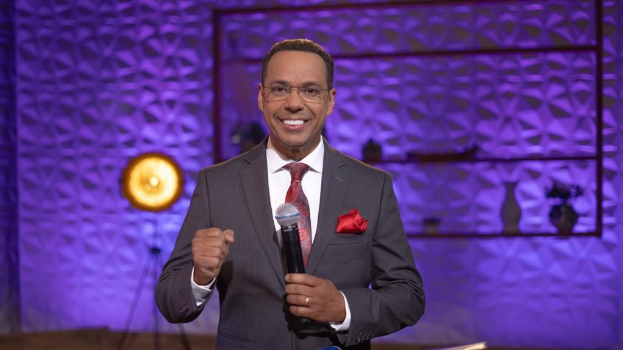 Stephen Colbert, Moderator von "The Daily Show with Trevor Noah", steht auf einer Bühne und hält ein Mikrofon in der Hand, trägt einen Anzug, Krawatte und Brille, während hinter ihm ein Scheinwerfer und ein Ständer zu sehen sind.