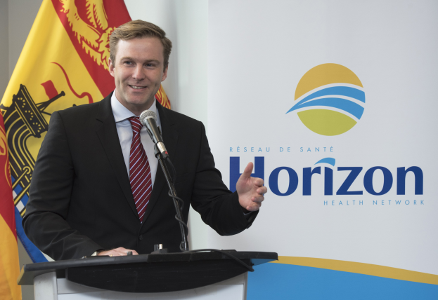 Ein Mann in einem schwarzen Blazer, weißem Hemd und roter Krawatte steht an einem Podium mit einem Mikrofon, lächelt und hat eine Fahne im Hintergrund.