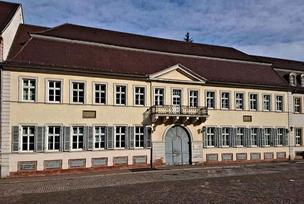 Ein großes Gebäude mit zahlreichen Fenstern, das Schlosshof in Baden-Württemberg, Deutschland, steht in der Mitte des Bildes, mit einer Straße unten und einem bewölkten Himmel im Hintergrund.
