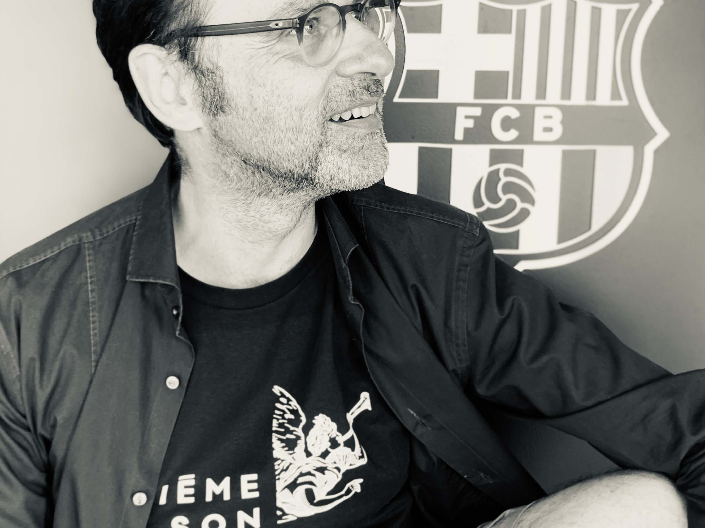 Ein Mann mit Brille und schwarzem Hemd sitzt vor einem schwarz-weißen FC Barcelona-Logo und sieht glücklich aus in einem klassischen Foto.