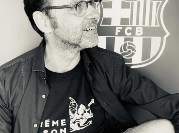 Ein Mann mit Brille und schwarzem Hemd sitzt vor einem schwarz-weißen FC Barcelona-Logo und sieht glücklich aus in einem klassischen Foto.