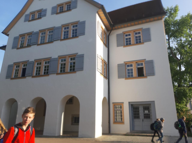 Eine Gruppe von Menschen, einige mit Taschen, geht vor einem großen weißen Gebäude mit Fenstern und Türen vorbei, umgeben von Bäumen unter einem klaren blauen Himmel, was darauf hindeutet, dass sie zu einer Schule in Deutschland gehen.
