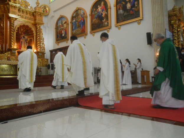 Eine Gruppe von Priestern, die einen roten Teppich in einer Kirche entlanggehen, mit Menschen, die vor und rechts von ihnen auf dem Boden stehen, und Foto Rahmen an der Hintergrundwand.