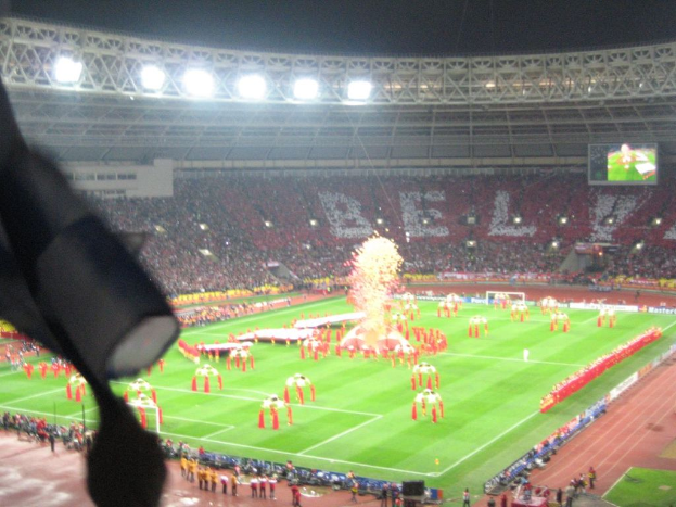 Ein Stadion mit Flutlicht, Eisen Gittern, Zuschauern, stehenden Personen, Werbetafeln, Anzeigetafeln, Überwachungskameras, Flaggen und Dekorationen.