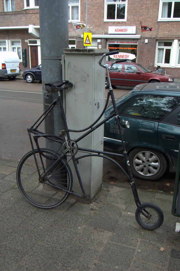 Ein Fahrrad steht neben Pfosten, Namensschildern, Fahrzeugen, Gebäuden mit Fenstern und Vorhängen sowie Fahnen.