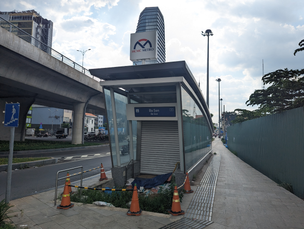 Eine Bushaltestelle an der Seite einer Straße neben einem hohen Gebäude, umgeben von Absperrbaken, Pflanzen, Gras, einem Zaun, Fahrzeugen, einer Tafel, Straßenlaternen, einer Brücke, Gebäuden mit Fenstern und einem bewölkten Himmel.