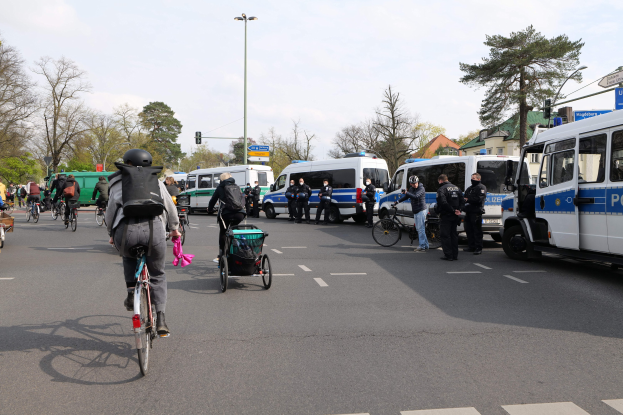 Eine Gruppe von Menschen, die auf Fahrr"adern eine Straäe entlangfahren, mit Polizeiwagen und anderen Fahrzeugen, einige tragen Helme und haben Taschen oder schieben Kinderwagen, vor einer Kulisse aus B"aumen, Geb"auden und Verkehrsinfrastruktur.