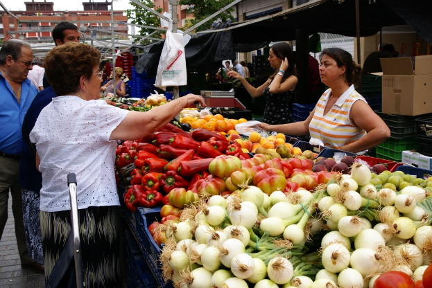 Gemüse in Kunststoffbehältern, eine Gruppe von Menschen, die stehen, Gebäude, Bäume, eine Kartonschachtel und der Himmel.