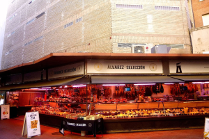 Ein belebter Markt mit verschiedenen Speisen, die von Deckenlampen beleuchtet werden und von Texttafeln umgeben sind, vor einem klaren blauen Himmel.