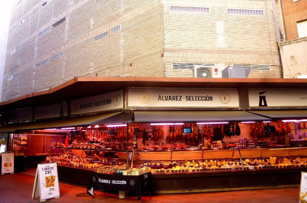 Ein belebter Markt mit verschiedenen Speisen, die von Deckenlampen beleuchtet werden und von Texttafeln umgeben sind, vor einem klaren blauen Himmel.