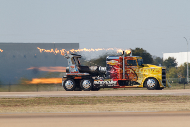 Ein Lkw mit Flammen, die aus seiner Rückseite kommen, fahrt entlang einer von Gras, einem Zaun, B├Ąumen, einem Geb├Ąude, einem Strommast, ├ťberlandleitungen und einem bew├Âlktem Himmel ges├Ąumten Stra├če, mit einem sichtbaren Reifen und Rad.