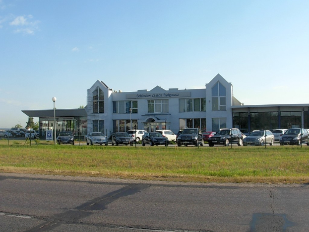 Ein Autohaus mit mehreren parkenden Fahrzeugen, umgeben von städtischer Infrastruktur wie Gebäuden, Straßenlaternen, Ampeln, fahrenden Autos, Infotafeln und Bäumen unter einem bewölkten Himmel.