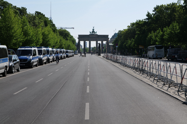Lange Reihe von Polizeiwagen auf der Seite einer Straße vor dem Brandenburger Tor in Berlin, Deutschland geparkt, mit Menschen auf Fahrrädern und auf der Straße stehend, Barrieren, Bäume, ein Bogen mit Statuen im Hintergrund und sichtbarem Himmel.