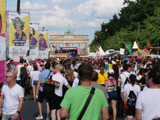 Eine große Menge Menschen, die eine Straße vor dem Brandenburger Tor in Berlin, Deutschland, entlanggehen, viele mit Kappen und Taschen, Banner links, sowie Bäume, Zelte, Ballons und ein Tor im Hintergrund bei einem bewölktem Himmel.