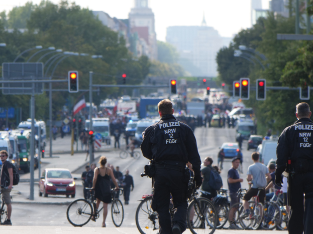 Zwei Polizeibeamte stehen an der Straßenseite neben einer Gruppe von Radfahrern, mit Fahrzeugen, Verkehrszeichen, Bäumen, Gebäuden und einem klaren blauen Himmel im Hintergrund.