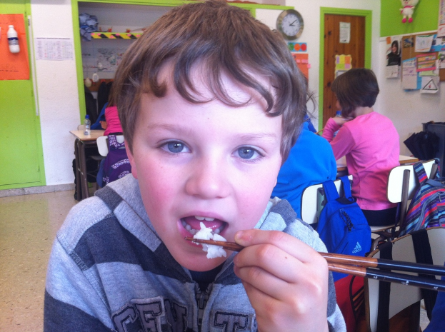Ein Junge in einer grauen und schwarzen Jacke isst mit Stäbchen in einem Klassenzimmer, mit anderen Kindern, die hinter ihm sitzen, einer grünen und weißen Wand, Lehrplänen und Postern an einer weißen Wand, einer braunen Tür und einer Wanduhr.