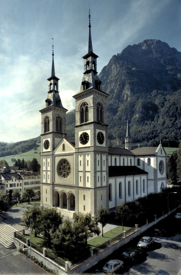 Luftaufnahme einer alten Kirche mit zwei Türmen in einer Stadt, umgeben von Gebäuden, Grünflächen, Fahrzeugen und Hügeln unter einem bewölkten Himmel.