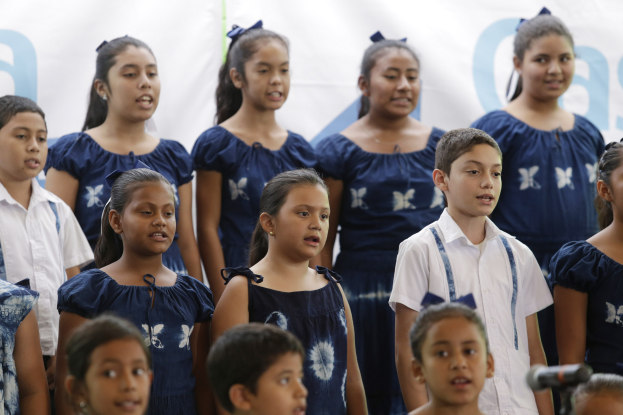 Eine Gruppe von Kindern steht vor einem Mikrofon und singt, mit einem Schulchor-Banner im Hintergrund.